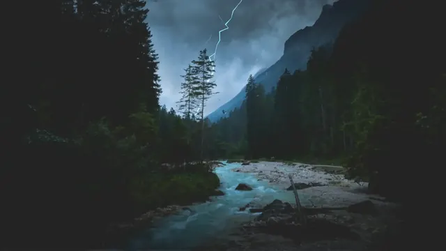 Lightning over a dark evergreen valley with a river and wet stones